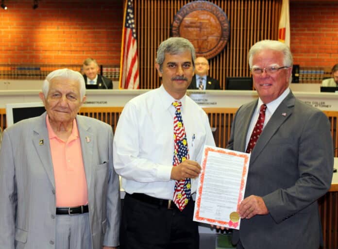 Dr. Singh receives Resolution from Commissioner Wayne Dukes, Second Vice Chairman, with Dr. Singh’s friend of 21 years, Mr. Nick Morana. Left to right:  Nick Morana, Pariksith Singh, MD, County Commissioner Wayne Dukes, Commission Chair Steve Champion look on from the Dais.