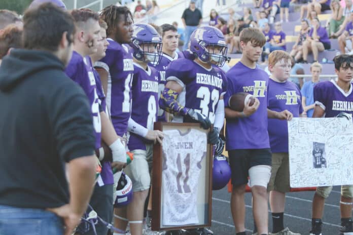 JV Football players gather around Blake Smith (8) and Nathaniel Greene (33) as they present Trevor's parents with his jersey and a poster created by those who knew and loved him during a half-time tribute to the late Trevor Bowen. 