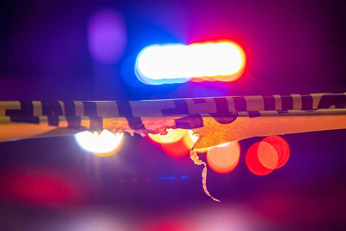 A defocused police car sits behind crime scene tape with flashing lights at night.